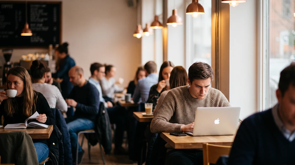 Person with focused expression using a laptop at a busy coffee shop with other patrons visible in the background, warm ambient lighting
