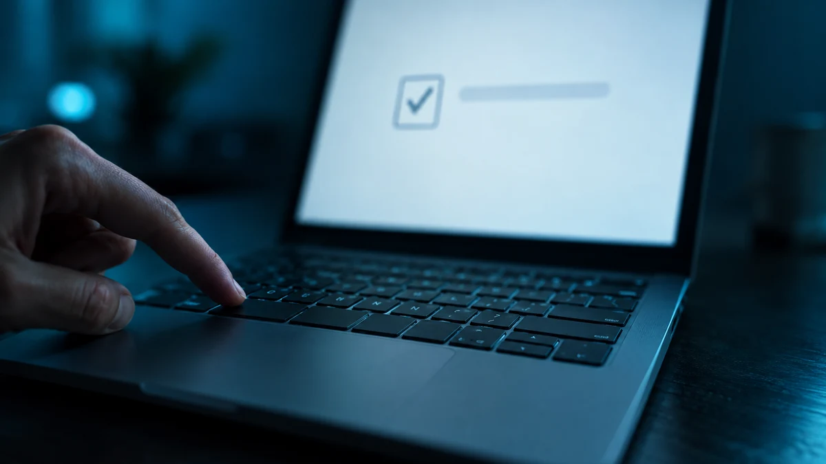 A hand resting near a laptop keyboard in a dim room while the screen shows a plain verification checkbox page, representing a fake CAPTCHA attack