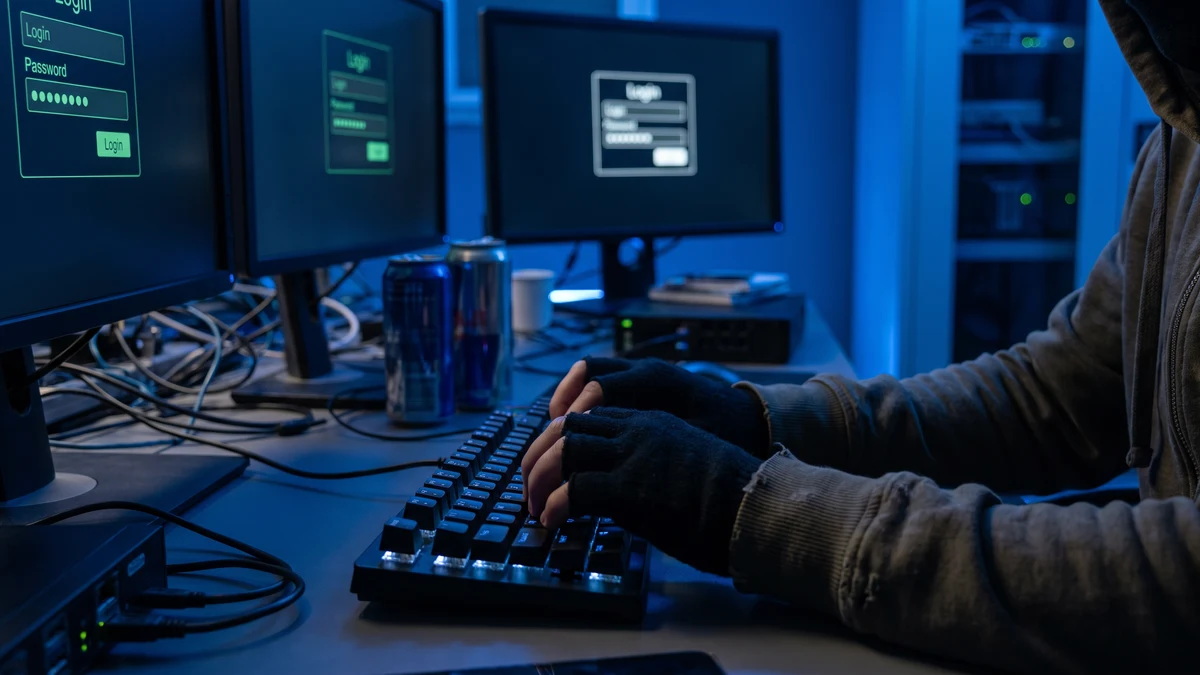 Hacker typing at keyboard in blue-lit environment with multiple login screens visible in background