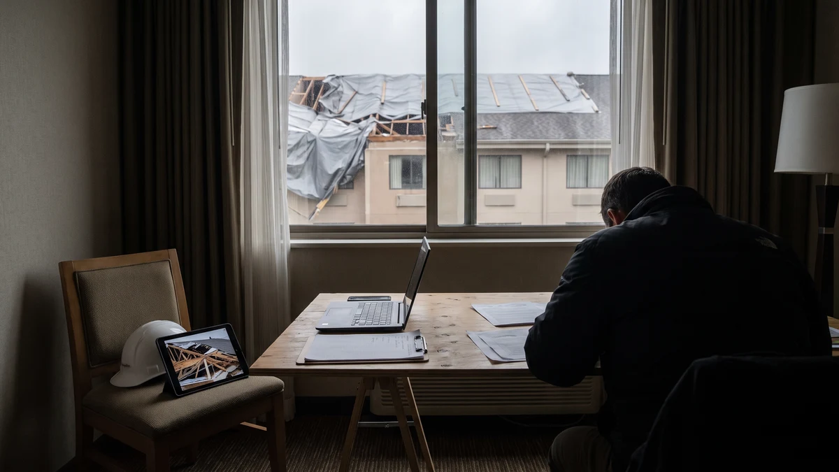 An insurance adjuster working on a laptop at a hotel room desk, with a storm-damaged roof visible through the window and a tablet showing damage photos